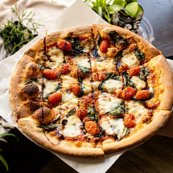 A hand holds a freshly baked pizza with cherry tomatoes, mozzarella, basil, and balsamic glaze.