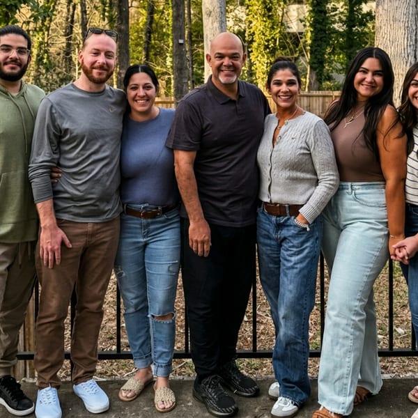 Seven smiling people, four women and three men, stand on a patio with a black railing and trees behind them.
