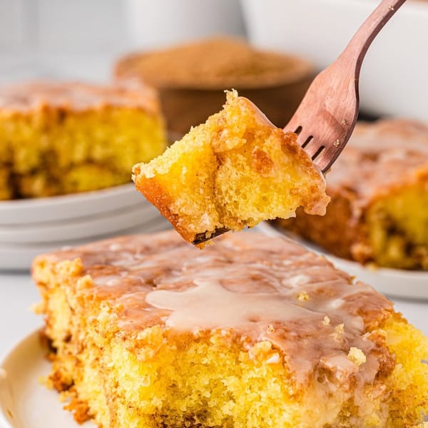 A fork lifts a bite of cinnamon roll cake with glaze, with more cake slices in the background.