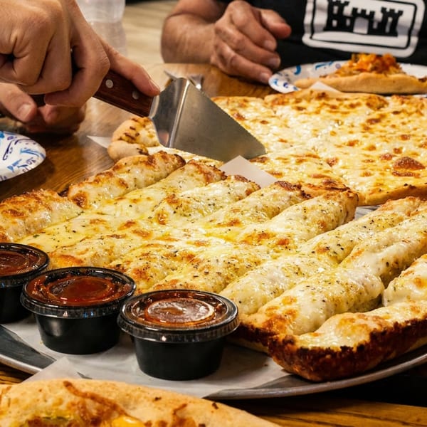 Hands serving cheesy breadsticks and a cheese pizza on a wooden table with dipping sauces.